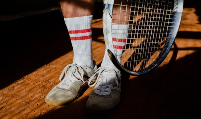 Close-up of tennis shoes, socks, and a racket on a clay court with shadows.